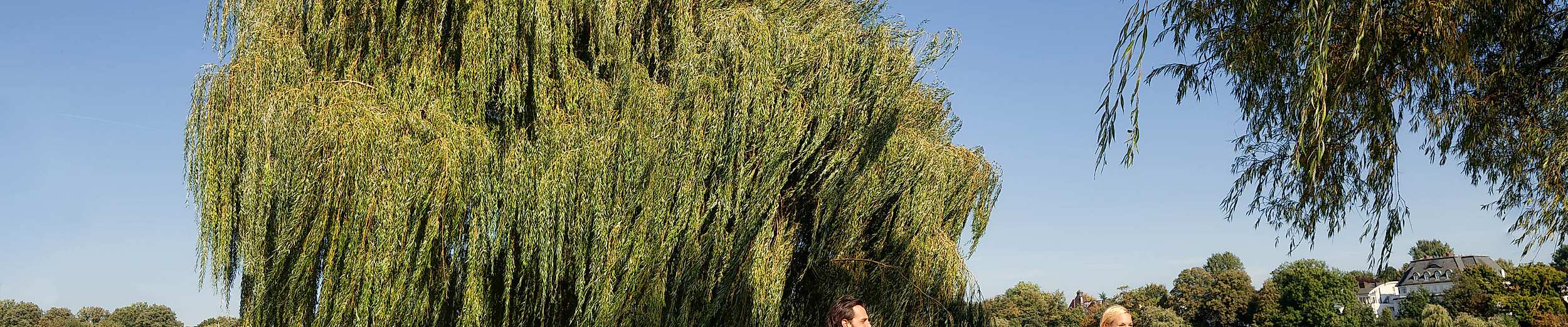 Zwei Personen radeln mit Stadträdern am Ufer der Alster entlang unter blauem Himmel.