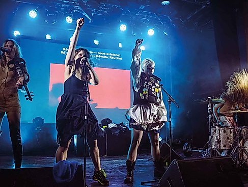 Three female performers are singing on stage, while a fourth is playing the drums.