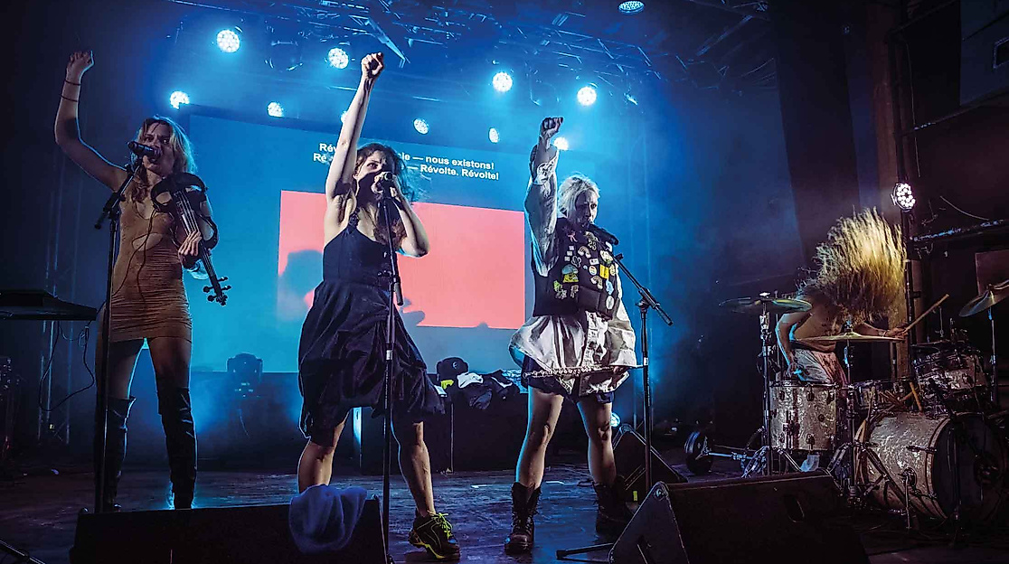 Three female performers are singing on stage, while a fourth is playing the drums.