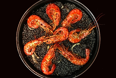 Pfanne mit schwarzer Paella und acht großen Garnelen auf dunklem Hintergrund stilvoll angerichtet.