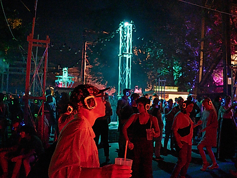 A nighttime JAJAJA event with a crowd wearing glowing red and blue silent disco headphones. In the foreground, a person wearing a cap and square glasses, holding a drink. In the background is an illuminated steel tower.