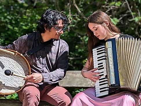 Víctor Gutiérrez and Isidora Caroca are sitting on a bench. While Víctor is holding a percussion instrument, Isidora is holding an accordion.