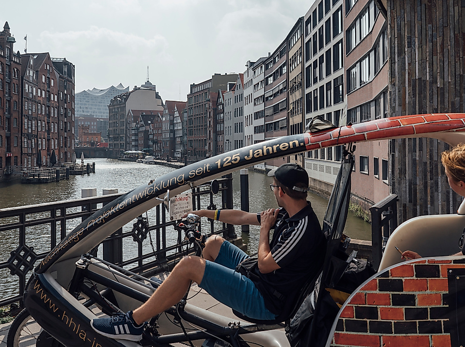 Zwei Personen fahren mit einer Rikscha durch die Hamburger Speicherstadt entlang eines Fleets.