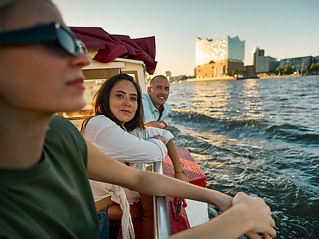 Three young people on a boat on the Elbe. The Elbphilharmonie concert hall is in the background.