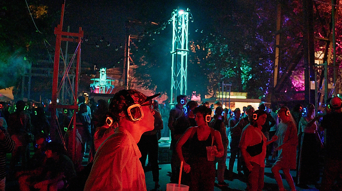 A nighttime JAJAJA event with a crowd wearing glowing red and blue silent disco headphones. In the foreground, a person wearing a cap and square glasses, holding a drink. In the background is an illuminated steel tower.