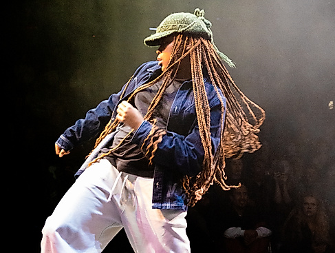 A person wearing a knitted hat, long braids, and loose clothing stands on a stage and moves their hips in one direction with bent knees. They pull themselves up in the same direction with the opposite arm.