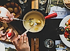 Geselliges Käsefondue mit Brot, Kartoffeln und Beilagen auf gedecktem Tisch, zwei Personen tauchen ein