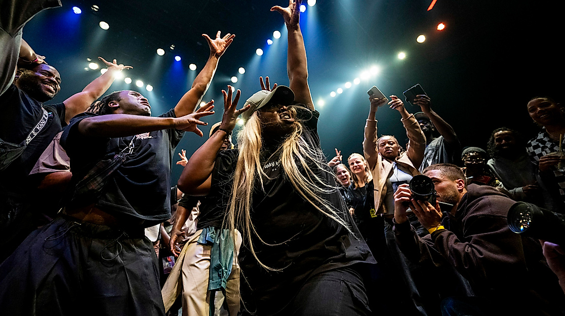 A dynamic scene in which a crowd of people stand around a dancer, cheering and cheering her on with gestures. The dancer raises her arms upwards, spreading her fingers. At the bottom right, someone photographs the dancer from close up.