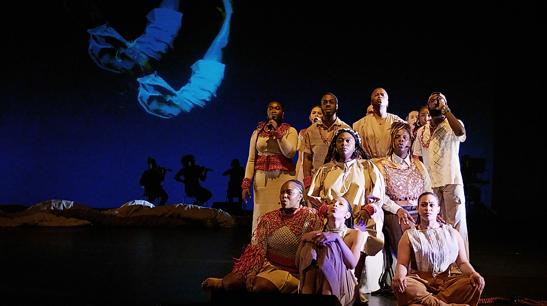 A group of performers in beige and red costumes sit and stand in formation on stage. In the background, silhouettes of musicians and a projection of a floating body are visible.