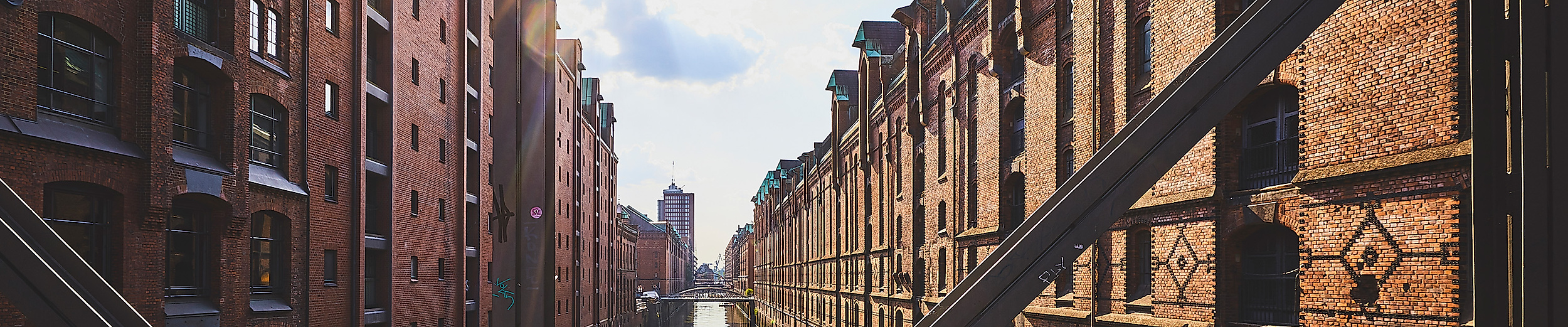 Blick durch eine Stahlbrücke auf die sonnenbeschienenen Backsteinfassaden der Speicherstadt entlang des Fleets