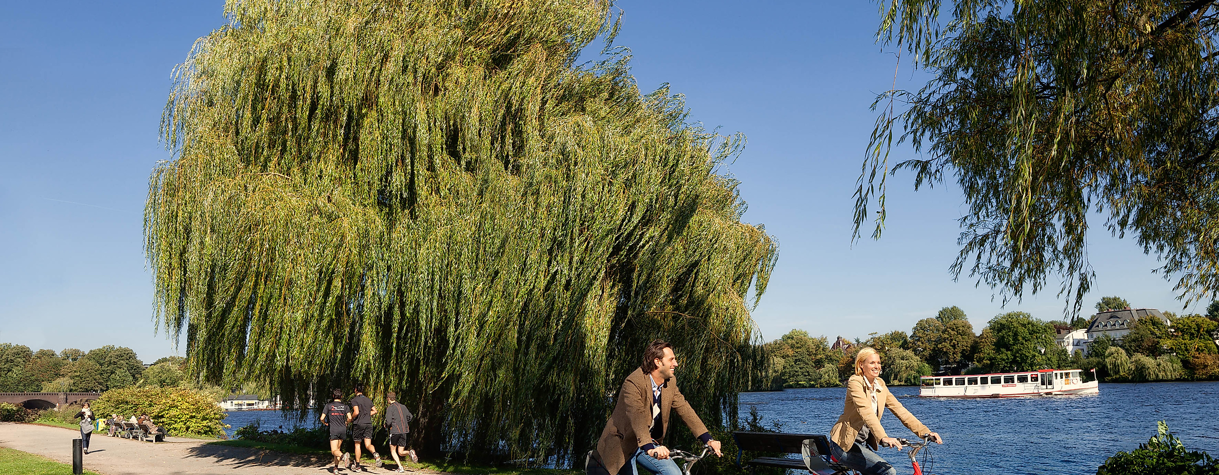 Zwei Personen radeln mit Stadträdern am Ufer der Alster entlang unter blauem Himmel.