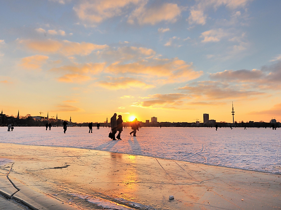 Menschen laufen Schlittschuh auf zugefrorener Alster bei Sonnenuntergang mit Blick auf Hamburgs Skyline
