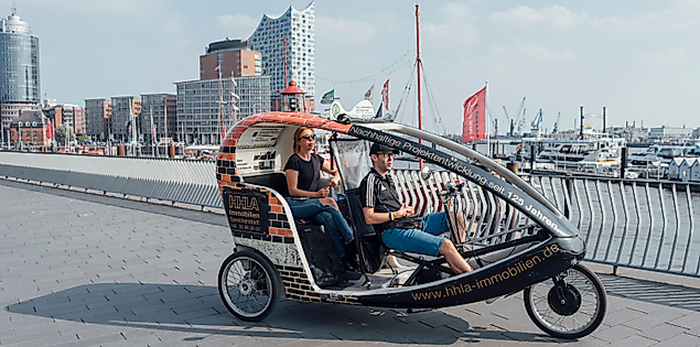 Rikscha mit zwei Fahrgästen an den Landungsbrücken, im Hintergrund die Elbphilharmonie