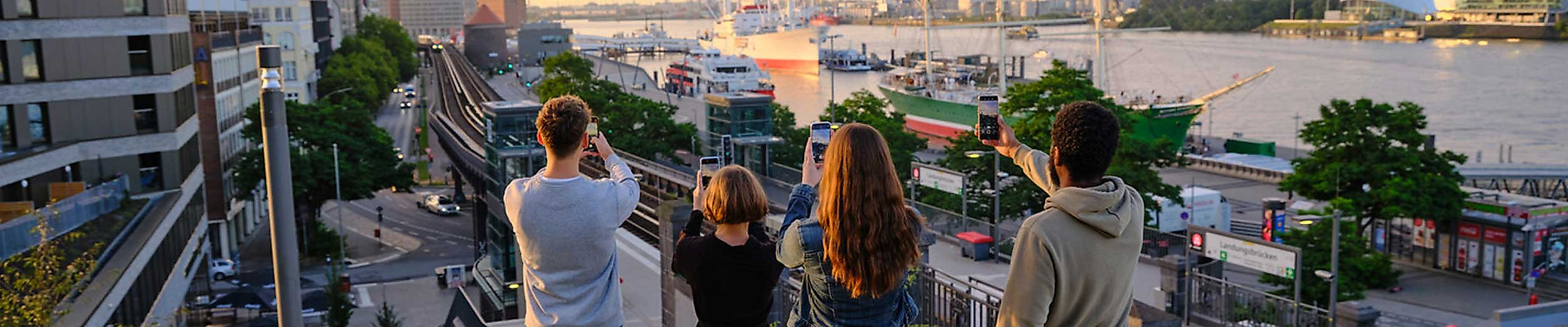 Ruckenansicht auf vier Erwachsene, die vom Stintfang Richtung Hamburger Hafen ein Foto mit Smartphone machen