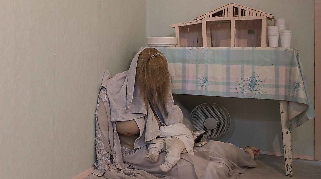 A person with long hair is sitting on the floor with a baby next to a table with a model house on it.