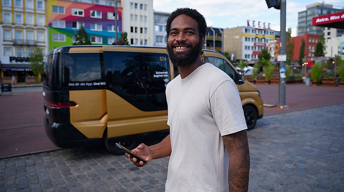 Mann mit Smartphone vor barrierefreiem goldfarbenem MOIA-Shuttle in urbaner Umgebung.