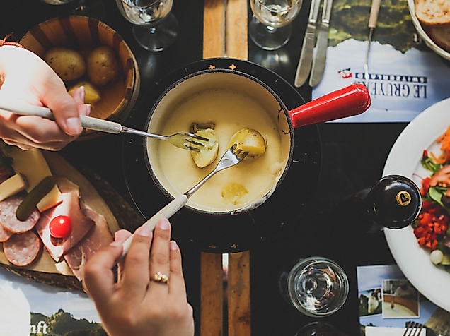 Geselliges Käsefondue mit Brot, Kartoffeln und Beilagen auf gedecktem Tisch, zwei Personen tauchen ein