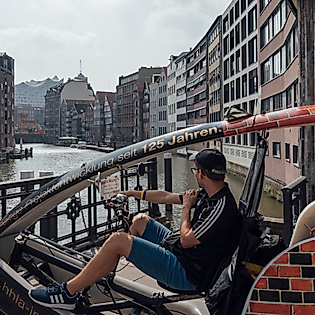 Zwei Personen fahren mit einer Rikscha durch die Hamburger Speicherstadt entlang eines Fleets.