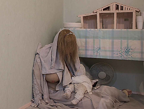 A person with long hair is sitting on the floor with a baby next to a table with a model house on it.