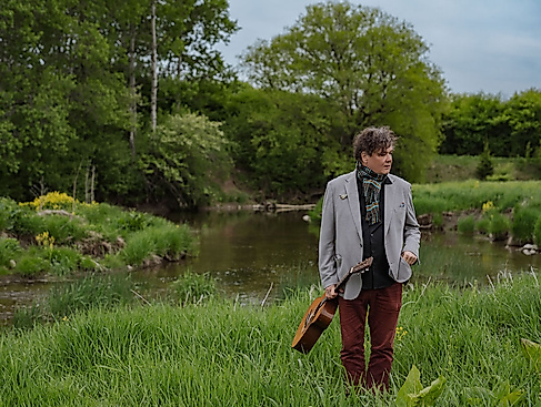 Ron Sexsmith stands in a green landscape holding his guitar.