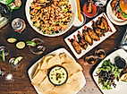 Große Auswahl an Speisen wie Salat, Chicken Wings, Hummus mit Brot und Burger auf Holztisch angerichtet.